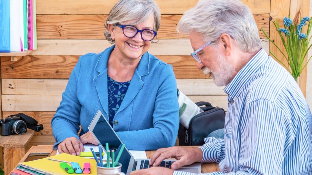 Cheerful couple of people with white hair sitting outdoor at a wooden table working with laptop computer.