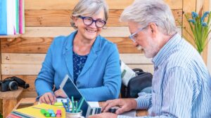 Cheerful couple of people with white hair sitting outdoor at a wooden table working with laptop computer.
