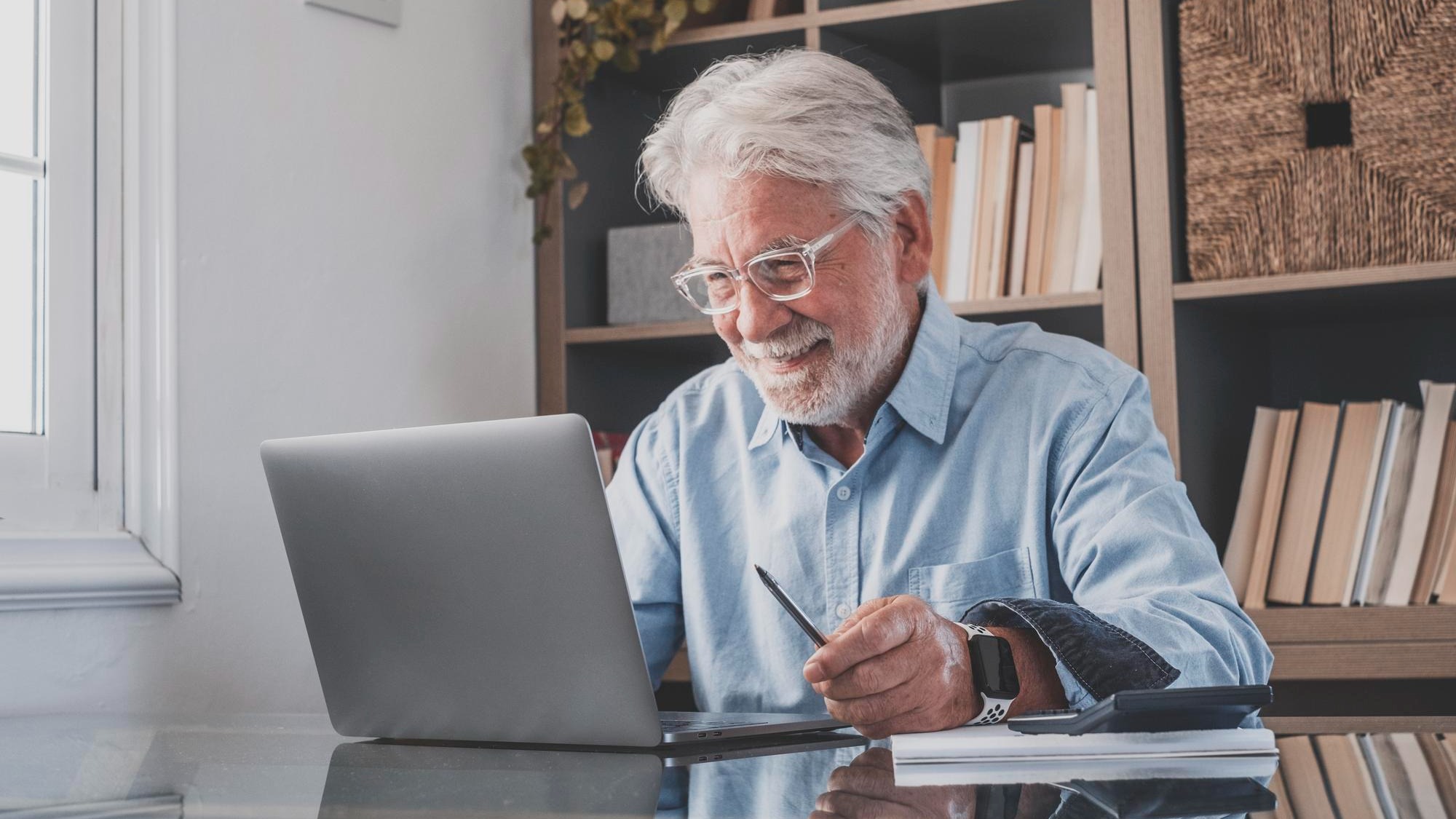 Happy old senior businessman looking at laptop screen and smiling on reading email sitting at home office Elderly man working online with calculator on table in front of book shelf at workplace Happy old senior businessman looking at laptop screen and smiling on reading email sitting at home office Elderly man working online with calculator on table in front of book shelf at workplace